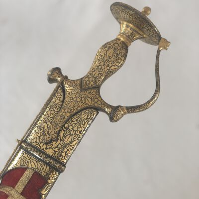 The image displays an ornate, decorative sword with a highly detailed golden handle featuring intricate, floral arabesque patterns and a red fabric-wrapped hilt, all set against a simple, light background, highlighting the craftsmanship and elegance of the weapon.