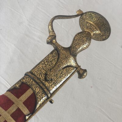 The image shows an ornate ceremonial sword placed on a white fabric background. This dagger features a highly decorated golden hilt with intricate floral patterns and a red fabric grip adorned with a cross pattern. The guard is similarly embellished, complementing the overall regal and antique appearance of the weapon.