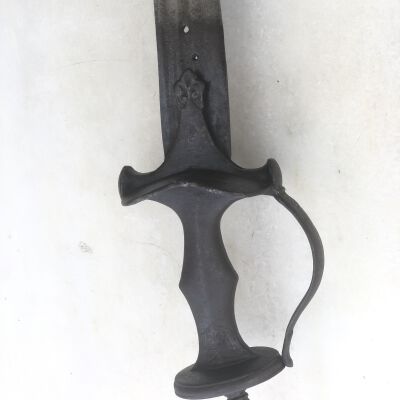 The image displays an aged, ornate sword against a pale, textured background. The blade appears long and somewhat tarnished, suggesting historical significance or perhaps neglect. The hilt is intricately designed, featuring a curved guard that loops around to protect the hand, adorned with floral designs and distinct textures that hint at its craftsmanship. The grip is smooth and dark, likely made from a hard material, shaped for a comfortable hold. A prominent pommel at the base complements the loop in the guard, adding balance and aesthetic appeal to the artifact. The overall condition and design of the sword point to its possible use in ceremonial contexts or as a decorative piece, reflecting the skills and artistic expressions of its era.
