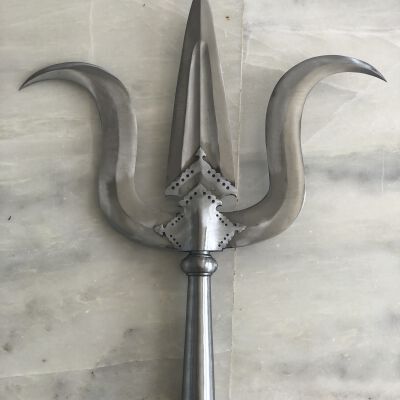 The image shows an ornate trident placed against a marble background. The weapon features a polished metal shaft topped with a triple-pronged head, each prong adorned with curving blades and detailed metalwork, giving it a charismatic and somewhat menacing appearance. The intricate design suggests that it could be ceremonial or a collector's piece, emphasizing craftsmanship and historical style.