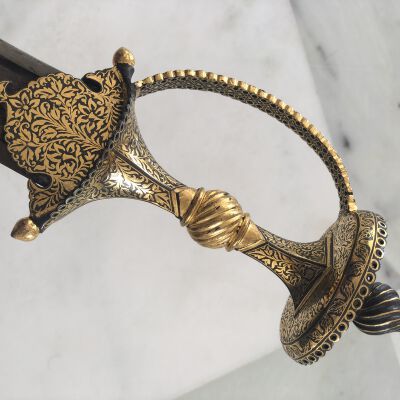 This image presents a meticulously crafted ceremonial dagger, featuring a distinctly ornate hilt richly adorned with intricate gold floral arabesque etchings on both the handle and the guard. The curvature of the guard adds a regal touch to the dagger, complementing its elegant swelling grip, which is also embellished with gold. Two prominent pommel-like structures, possibly serving decorative purposes, can be seen on either end of the guard, further enhancing the sword's aesthetic. The blade itself is not completely visible, suggesting the focus of the image is primarily on the elaborate craftsmanship of the hilt and its components. The sword is photographed against a subtly textured gray marble background that contrasts with the elaborate gold detail, highlighting the craftsmanship and grandeur of the piece. This sword likely serves as more of a ceremonial or decorative artifact rather than a functional weapon, emphasizing status, power, and artistry in its design and execution.