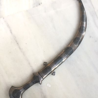 The image displays a sheathed Jambiya dagger lying on a pale marble surface. The dagger features a distinctive curved shape, and the sheath and handle are adorned with intricate silver floral designs. Each section of the sheath is highlighted with darkened backgrounds that enhance the silvery patterns, and the handle has decorative elements near its widening base, emphasizing traditional craftsmanship. The decorative artwork is finely detailed, suggesting a blend of utilitarian and artistic value typical of such historical pieces.