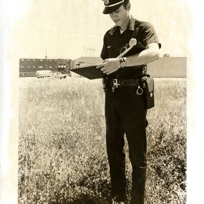 Policeman standing in field, writing in binder: "Another violation"