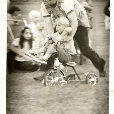 Man pushing boy on tricycle- spectators: Broomfield Days?