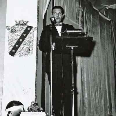A B/W photograph of a man speaking at a podium.