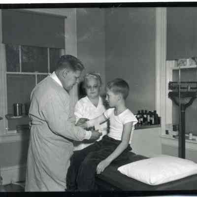 Dr. bandaging the arm of a young boy with nurse watching.