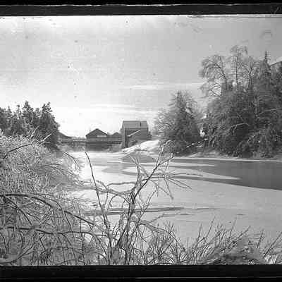 An alternate view of the bridge and mills: The mills visible in this picture include, right to left, the bone and grist mill, the saw mill, and the corner of the box and novelty mill. Water pours from the sluice running under the right end of the Upper Bridge, while an ice cap teeters on the large rock associated with John James Audubon in the middle of the river.