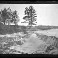          Water floods over Lincoln Dam on the Dennys River, c. 1885; View of spring floodwater rushing over the Dennys River dam in the springtime, with shaft across the flowage in the foreground to one of the mills.   The State Seal Pines are clearly visible on the Edmunds side of the river.
   