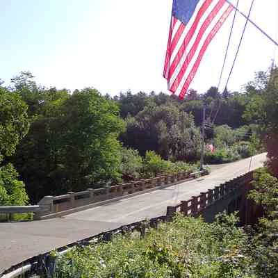 Upper Bridge over the Dennys River in 2023: The Upper Bridge photographed in the June sunshine of a midsummer's morning decorated with flags in preparation for the Fourth of July parade in a few weeks' time.   The River Road in Edmunds joins the Bunker Hill Road as it heads up the hill towards the start of the parade route at the Edmunds Consolidated School.