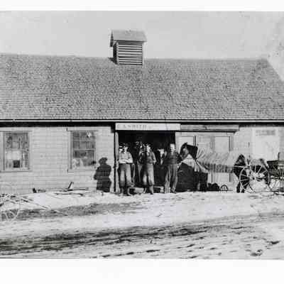 Charles A. Smith's blacksmith shop, next to his house on the River Road in Edmunds, Maine: "Chick" Smith is to the right of the group of men standing in the doorway of his blacksmith shop, where a pair of overalls covers his wooden leg.