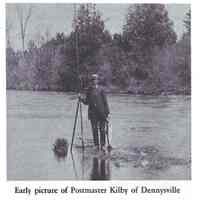          Howard Kilby, Dennysville Postmaster displaying a freshly caught salmon caught on the Dennys River in the late nineteenth century.; Note the length of his fly rod, waders and the gaff hook in his hand, as well as the size of his catch.
   
