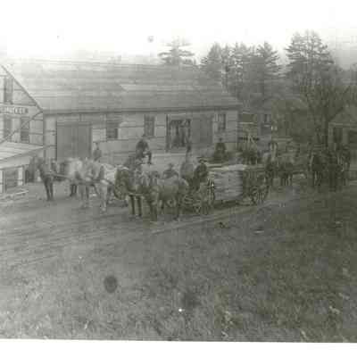 Dennysville Lumber Company, Dennysville, Maine: Teamsters and their loads prepare to transport lumber to market from the Dennysville Lumber Company, rebuilt after a fire in 1907.  The small Company office building and the rooves of several other mills on the Dennys River are in the background, with the State Seal Pines on the Edmunds side of the river beyond.