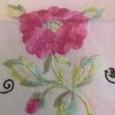 The image shows an intricately embroidered pink flower with green leaves on a light fabric background.